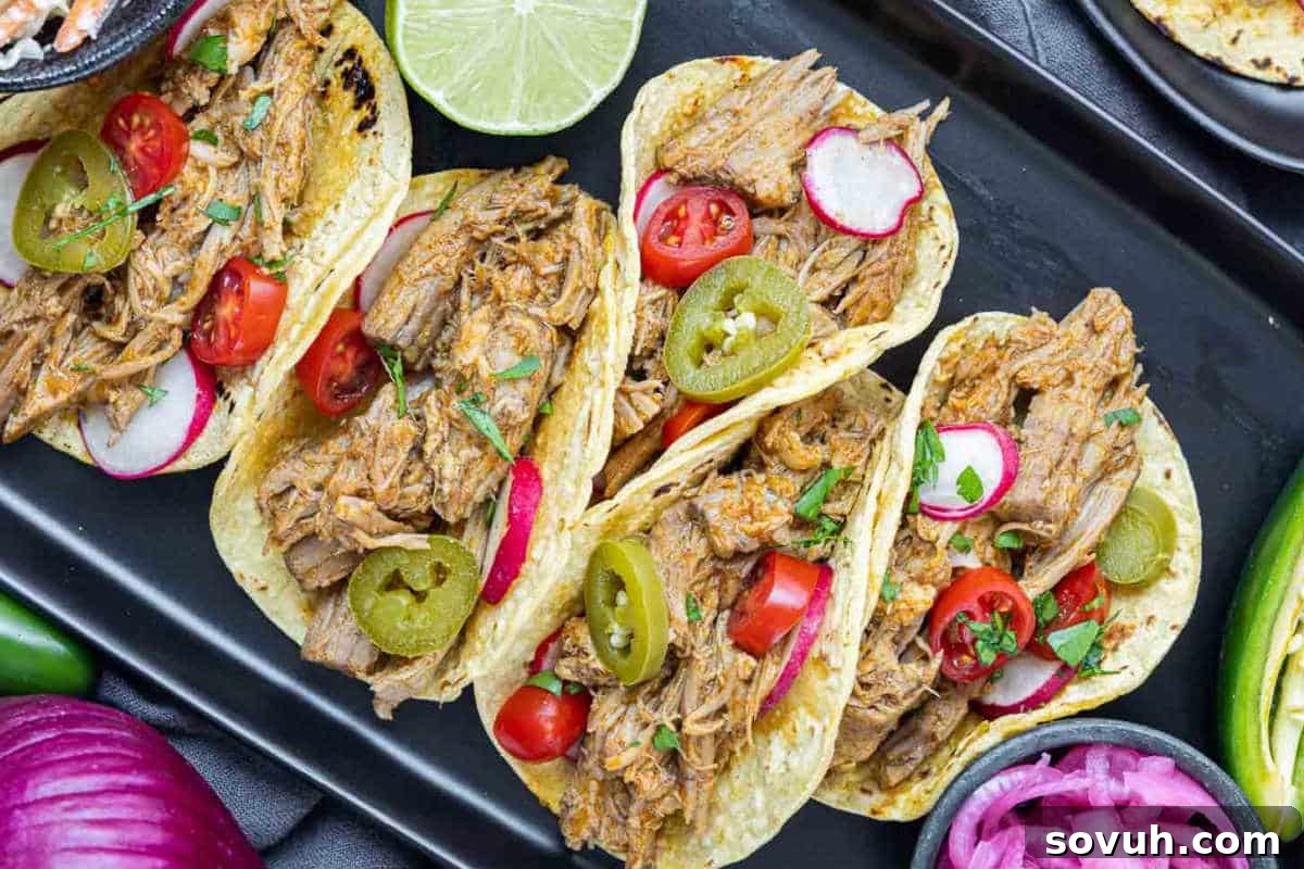 Four tacos filled with shredded meat, garnished with sliced radishes, jalapenos, cherry tomatoes, and cilantro, served on a black tray with a lime half and colorful vegetables around.