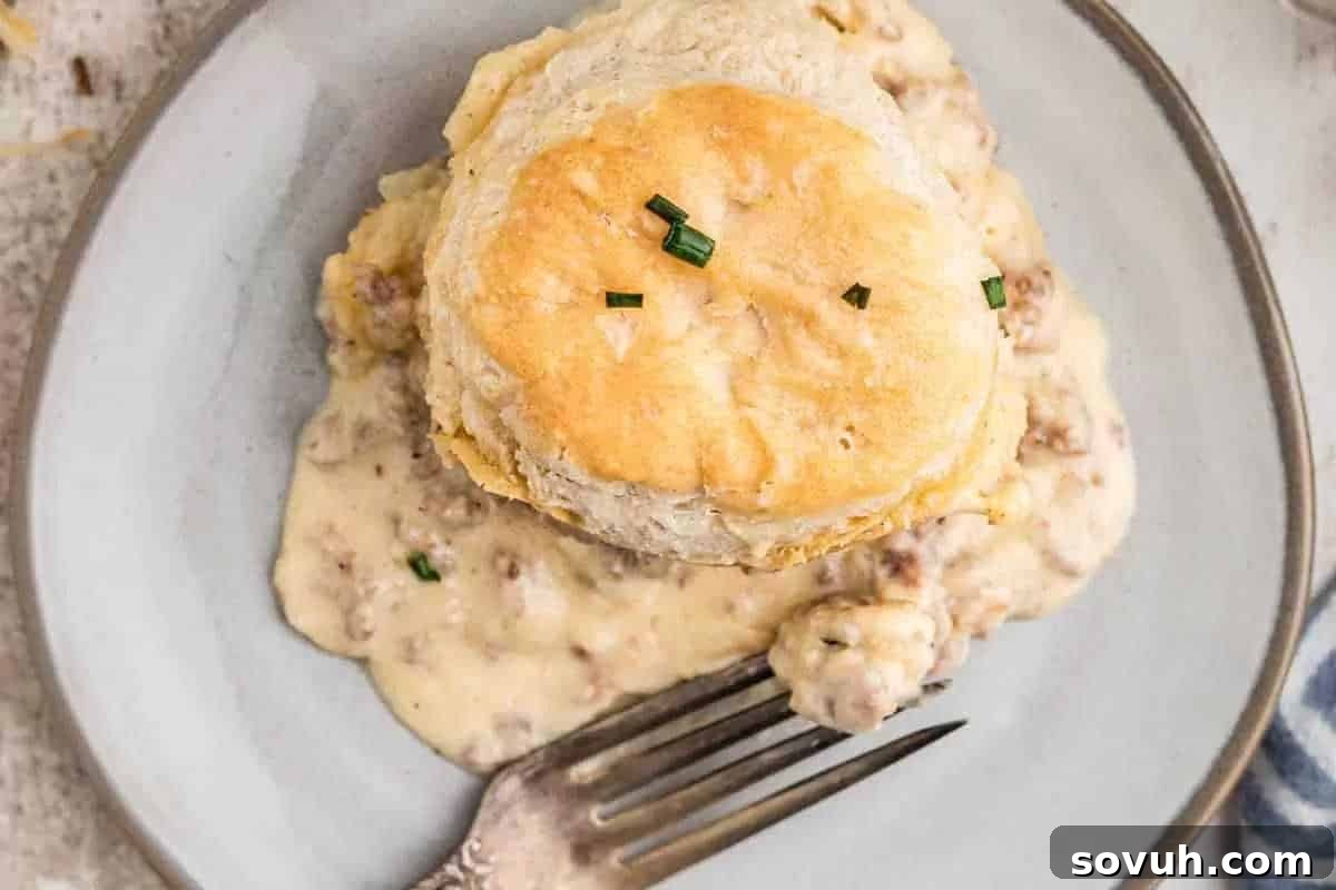 A biscuit topped with chopped chives sits on a plate with sausage gravy beside a fork.
