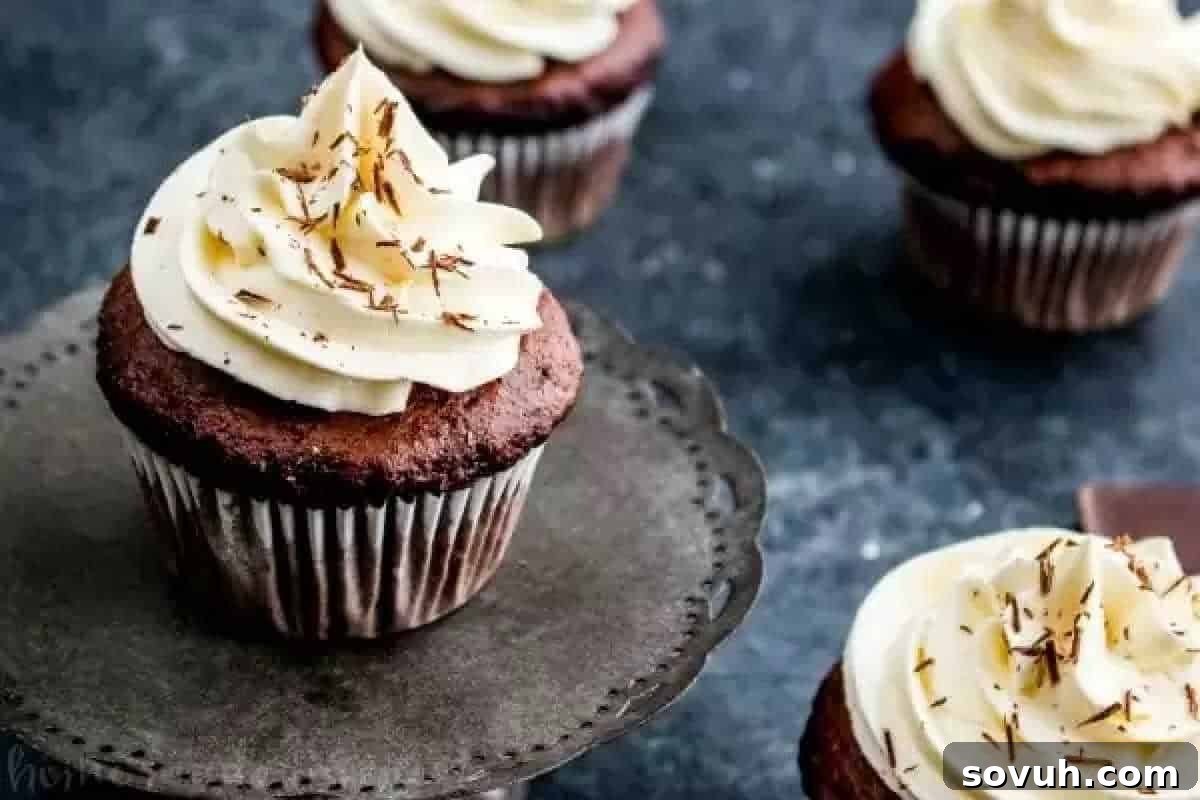 Cake Mix Magic 2 Four chocolate cupcakes with white frosting and chocolate shavings on top, placed on a dark surface. One cupcake is on a small metal stand, showcasing delightful desserts for those who appreciate Sweet Cheats.
