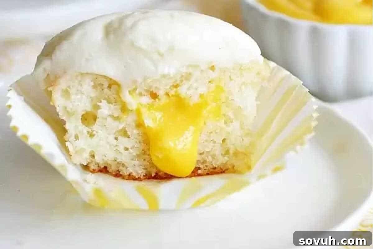 Cake Mix Magic 16 A cupcake with white frosting and yellow filling, partially eaten, sits on a plate with its paper liner peeled back. A small bowl with yellow filling is visible in the background.