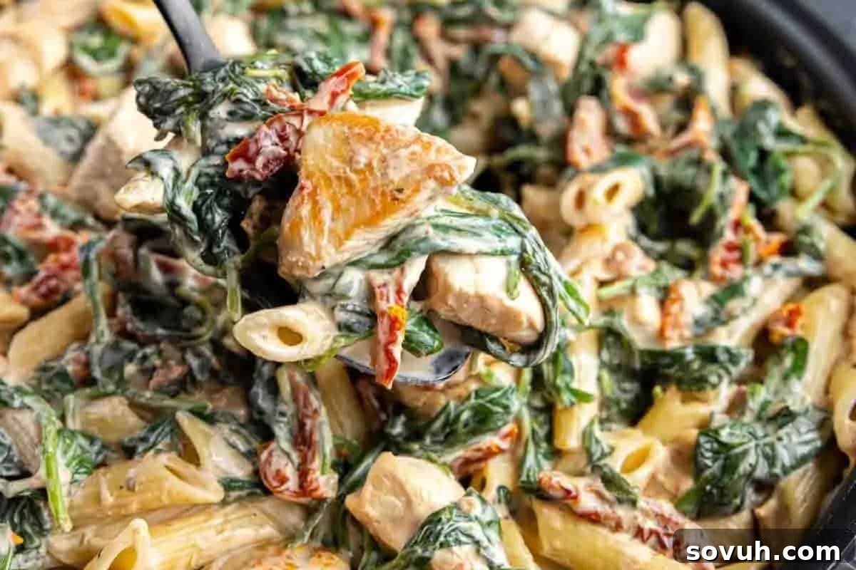 A close-up of a creamy and vibrant pasta dish, featuring penne pasta, tender chicken pieces, fresh spinach, and sun-dried tomatoes, being gracefully served with a spoon. The rich sauce coats every ingredient beautifully.