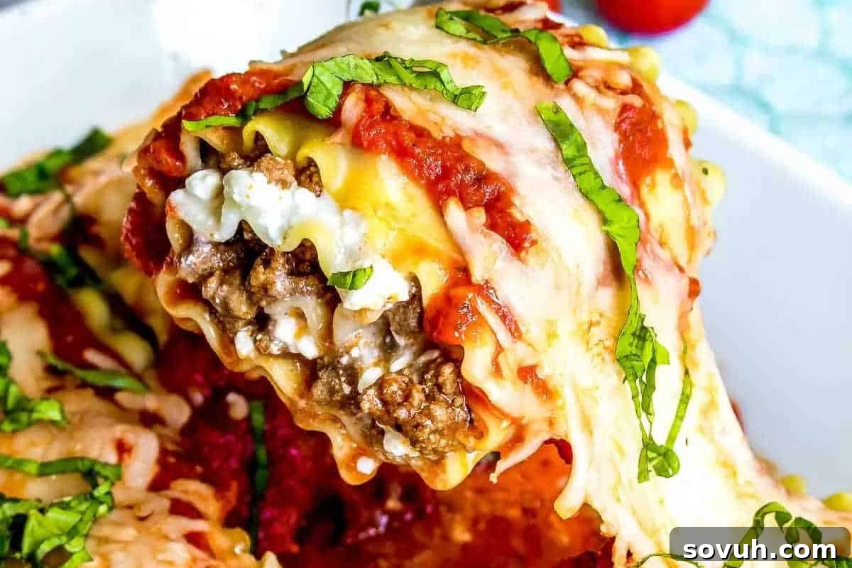 A close-up of a delectable cheesy lasagna roll-up being lifted from a white baking dish. The roll reveals layers of tender pasta, savory ground meat, creamy ricotta cheese, and rich tomato sauce, all crowned with melted cheese and fresh basil ribbons.