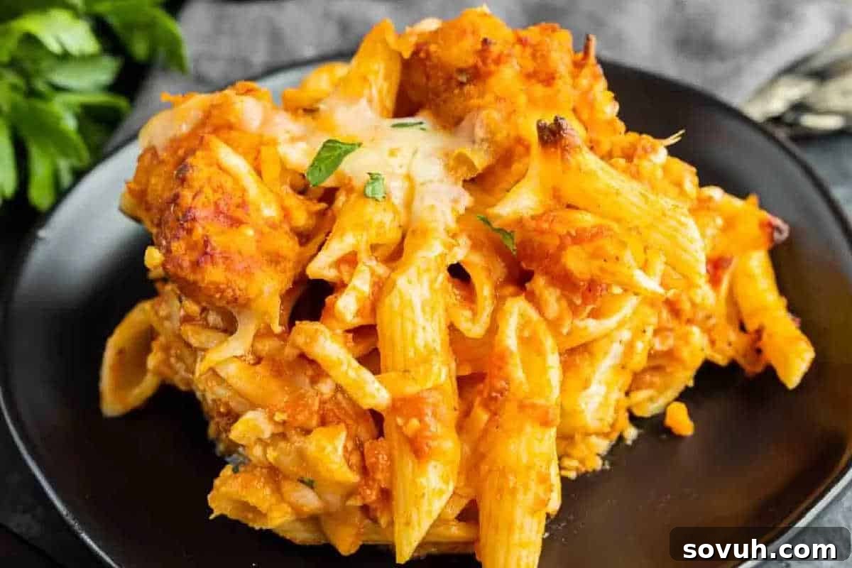 A close-up shot of a serving of baked penne pasta with chicken parmesan on a black plate, artfully garnished with chopped fresh herbs. The dish features a delectable golden-brown melted cheese crust and a rich, inviting tomato sauce.