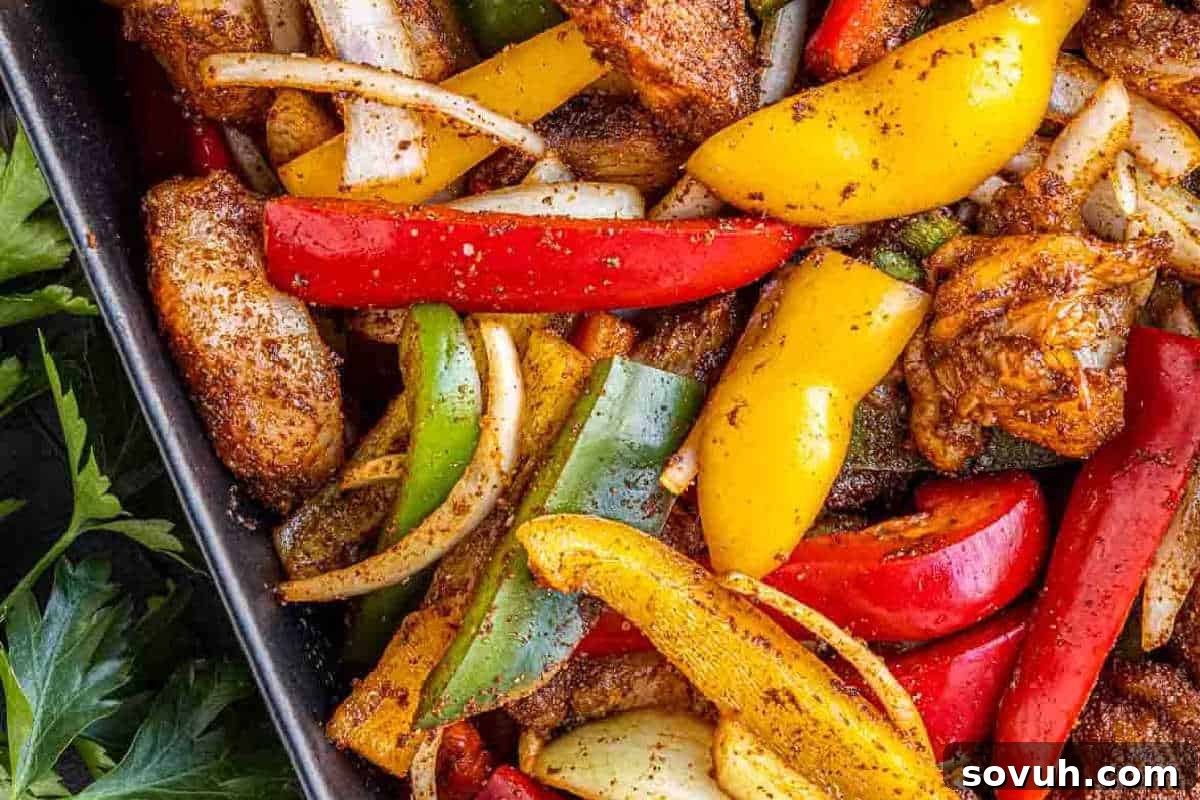 A vibrant close-up of a sizzling sheet pan filled with colorful roasted vegetables and perfectly seasoned chicken strips, including bright red, yellow, and green bell peppers, sweet onions, and tender chicken pieces. Fresh parsley adds a pop of green on the side.