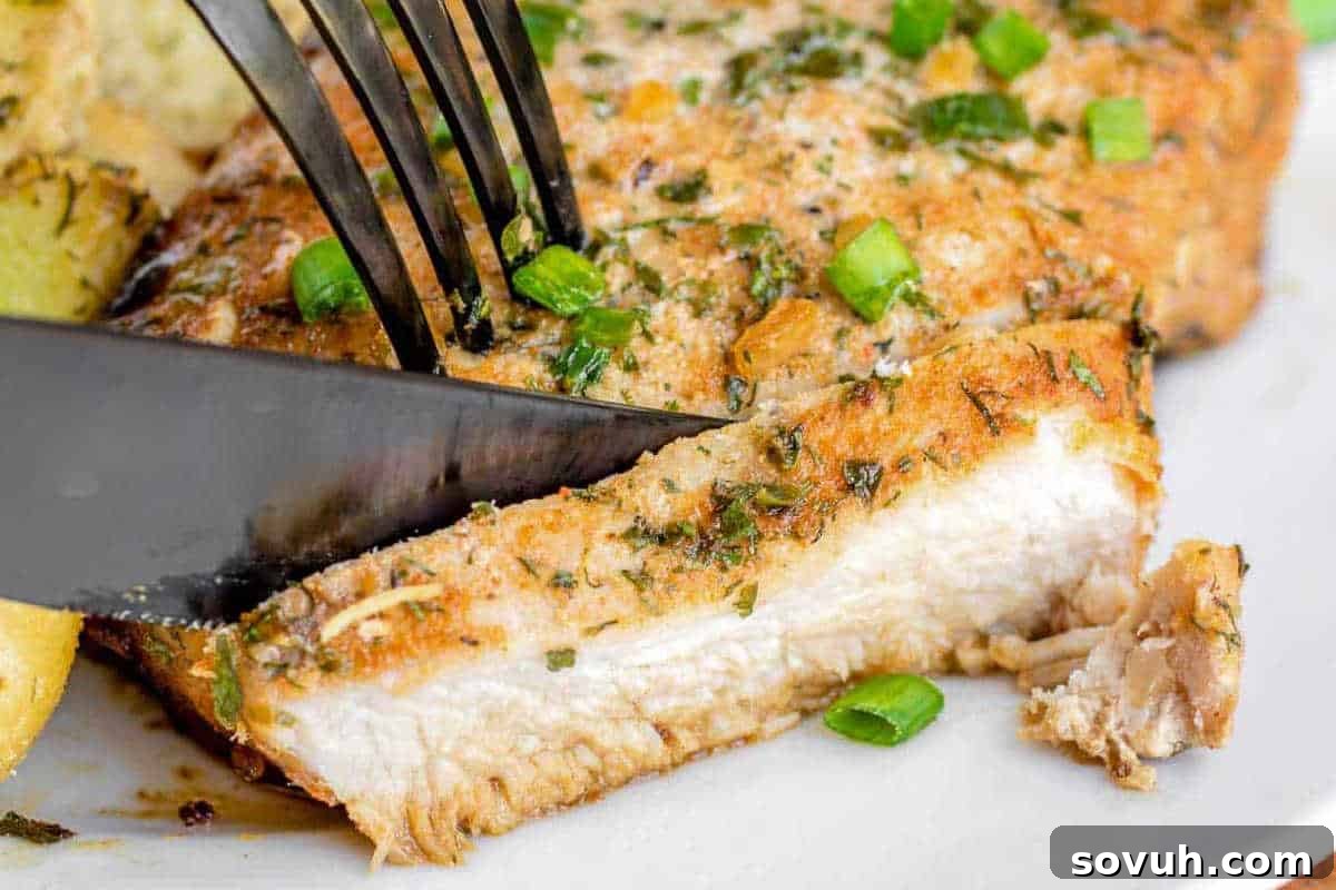 Succulent baked ranch pork chop, perfectly seasoned, resting on a plate with a fork and knife, ready to be enjoyed. Garnished with fresh green onions and a side of roasted potatoes.