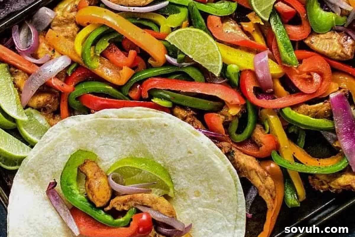 A warm tortilla piled high with perfectly seasoned chicken and vibrant strips of colorful bell peppers, resting on the edge of a baking tray filled with more cooked chicken, bell peppers, and onions, with fresh lime wedges adding a zesty touch.