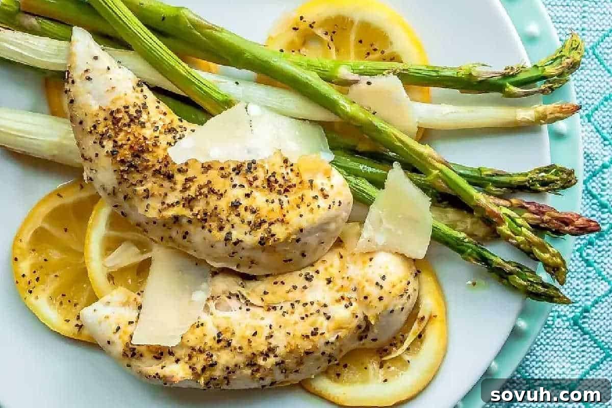 Grilled chicken breasts seasoned with freshly ground pepper and melted cheese, elegantly served with bright green asparagus spears and vibrant lemon slices on a pristine white plate, making a healthy and appealing meal.