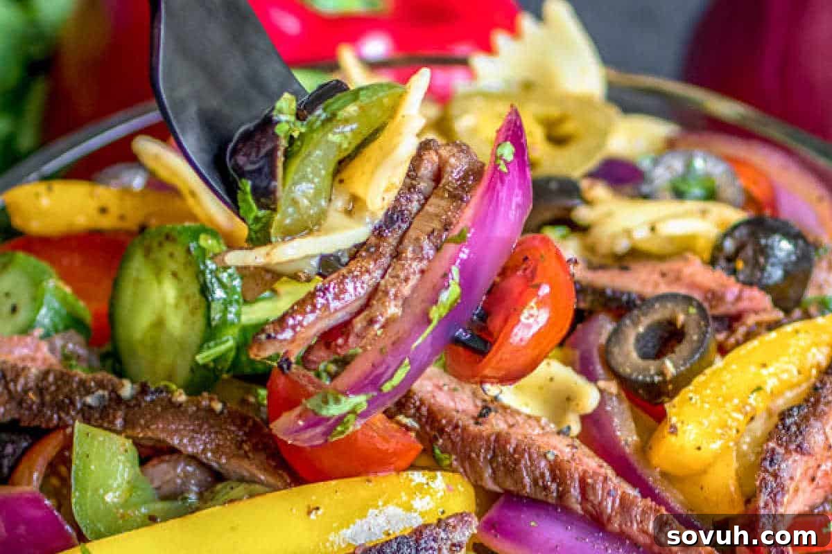 A close-up shot of a fork holding a mix of pasta, grilled vegetables, and sliced beef from a colorful and fresh pasta salad.