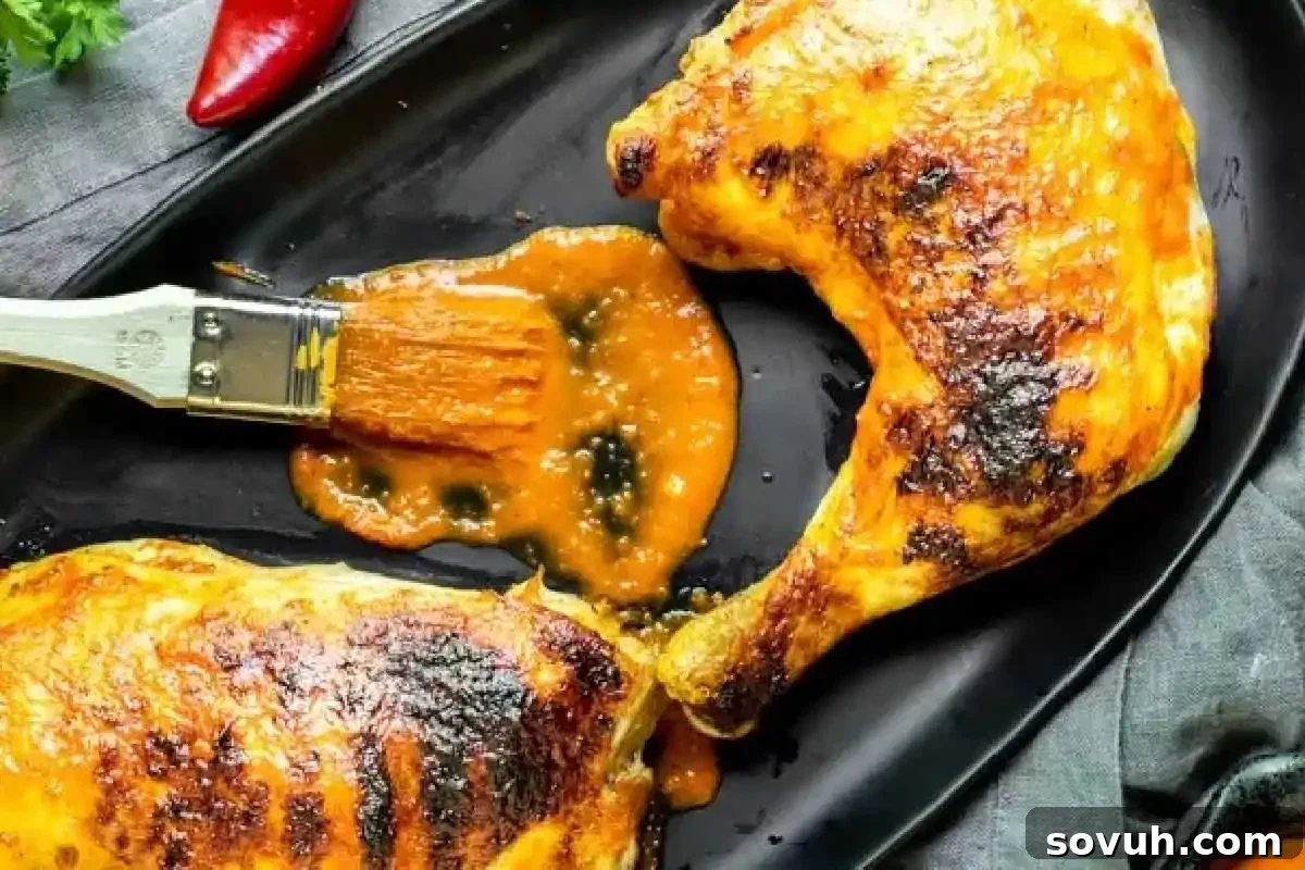 Grilled chicken legs with golden-brown skin on a black tray, one being brushed with orange sauce. A red chili and parsley are in the background.