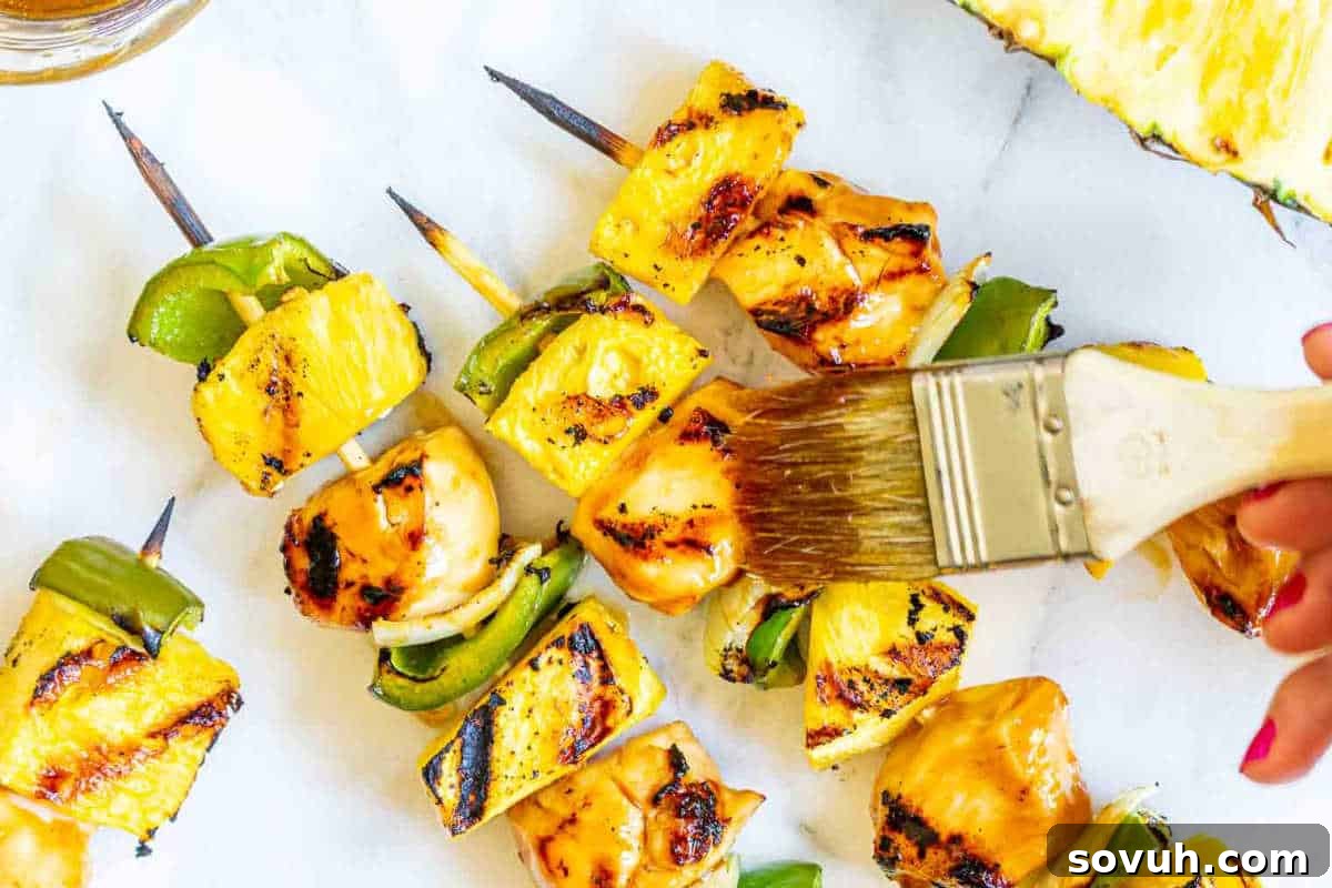 Grilled pineapple and chicken skewers with green bell peppers are being brushed with marinade.