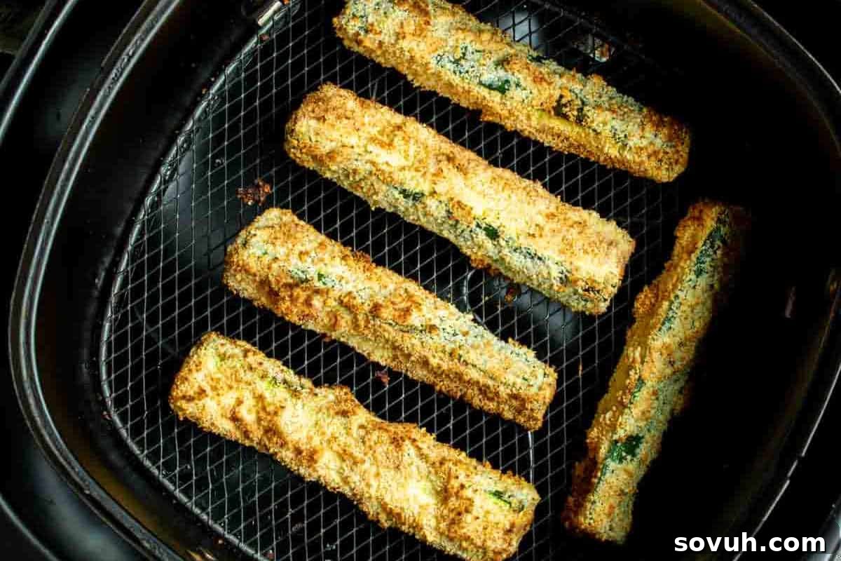 Five breaded zucchini sticks are placed on a black wire rack, cooking inside an air fryer.