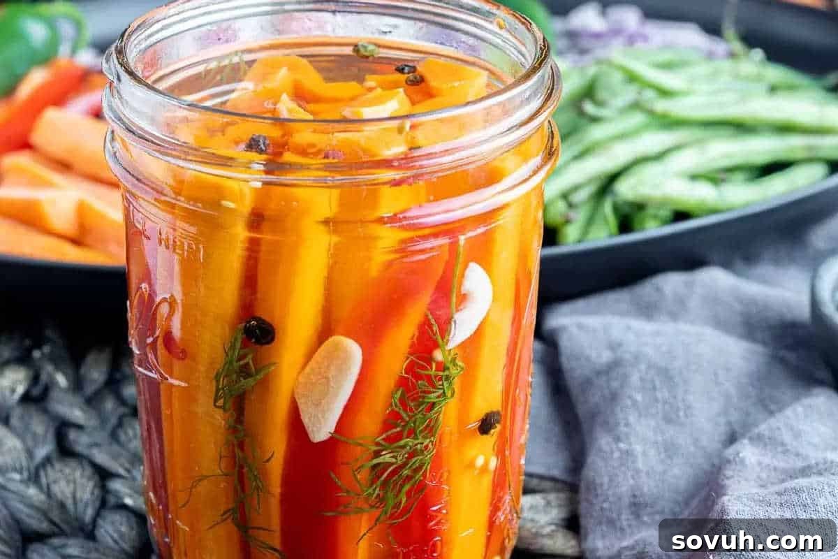 A mason jar filled with pickled vegetables including carrot sticks and red bell pepper slices, garnished with garlic cloves, dill, and peppercorns. A plate of fresh green beans is in the background.