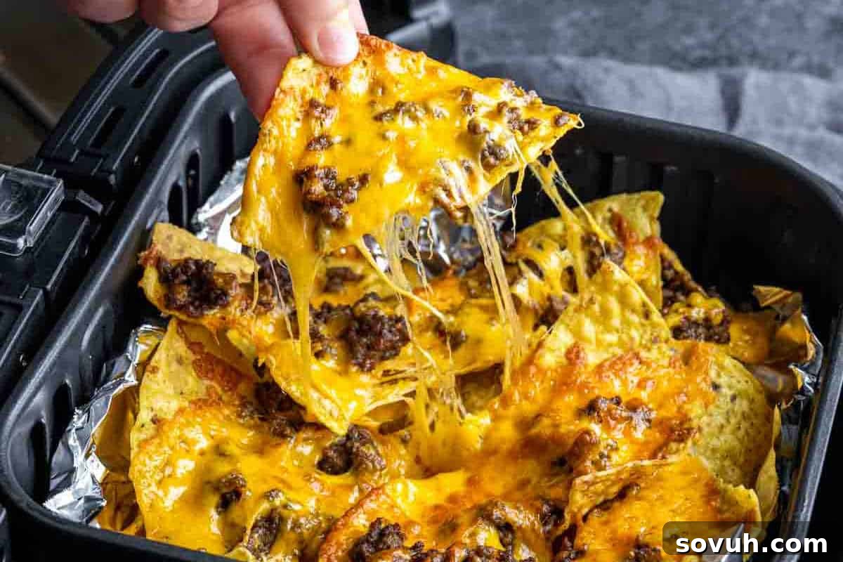 Close-up of a hand lifting a cheesy nacho with ground beef from a tray of nachos in an air fryer, with melted cheese stretching from the tray to the nacho.