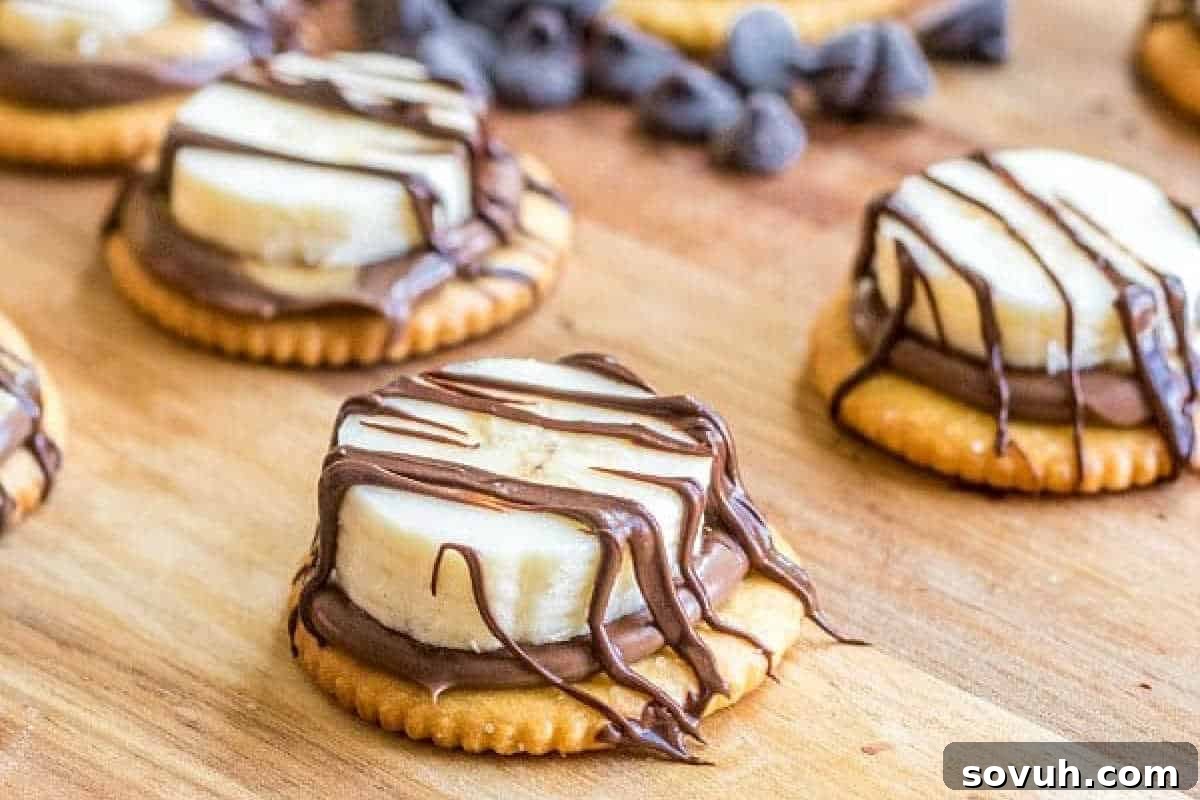Crackers topped with chocolate spread, banana slices, and drizzled with chocolate sit on a wooden surface.