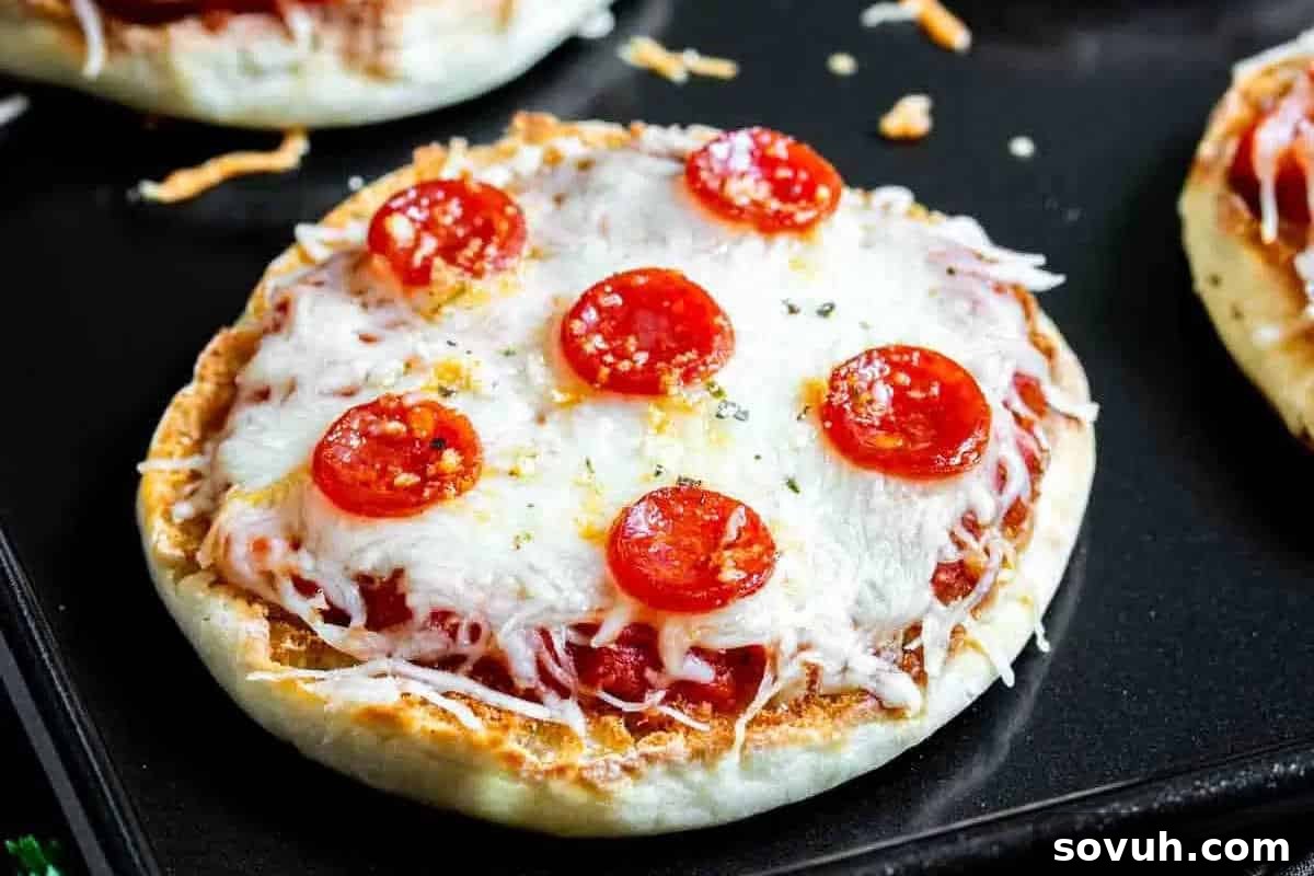 A mini pizza topped with melted cheese, slices of pepperoni, and a sprinkle of seasoning is placed on a black baking tray.