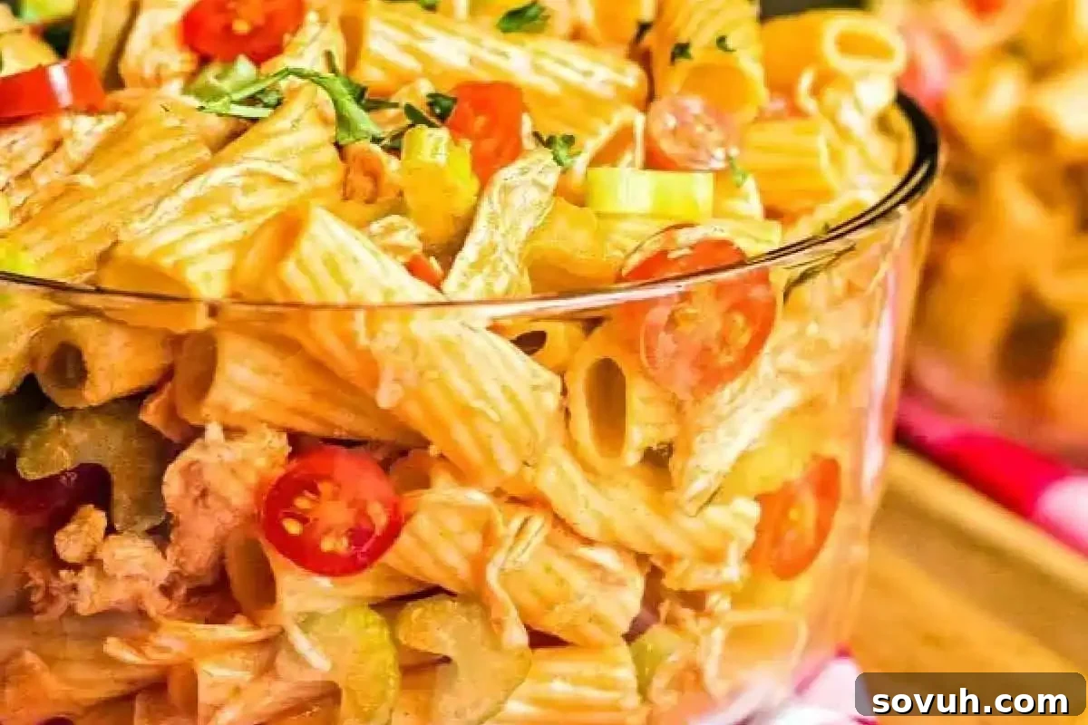 A clear bowl filled with creamy pasta salad, featuring rigatoni noodles, sliced cherry tomatoes, and celery pieces, garnished with herbs.