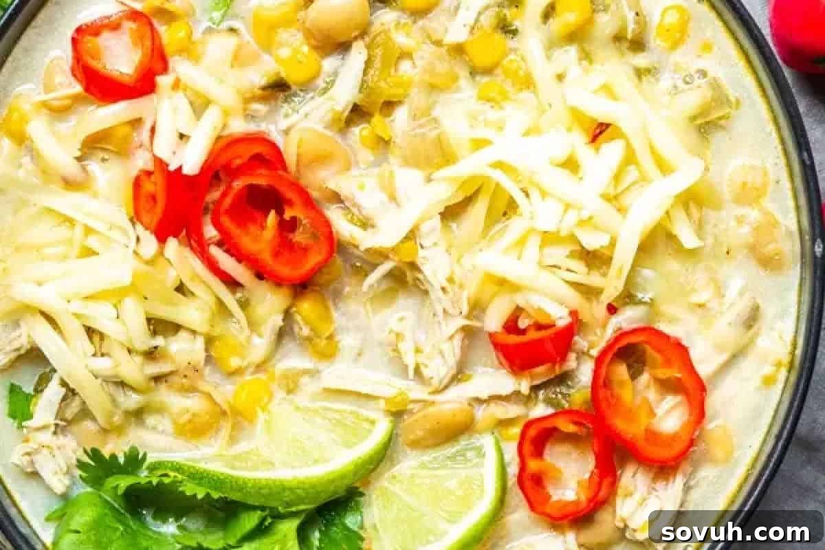 A bowl of creamy chicken chili is topped with shredded cheese, sliced red peppers, lime wedges, and cilantro. Corn and beans are visible among the ingredients.