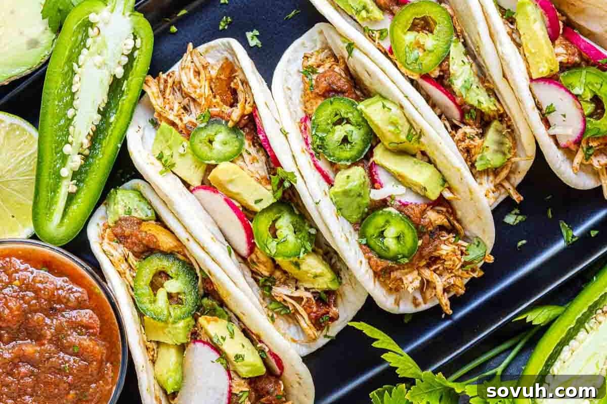 A tray of tacos filled with shredded meat, sliced avocado, jalapeño, radish, and garnished with herbs, surrounded by lime wedges and a bowl of salsa.