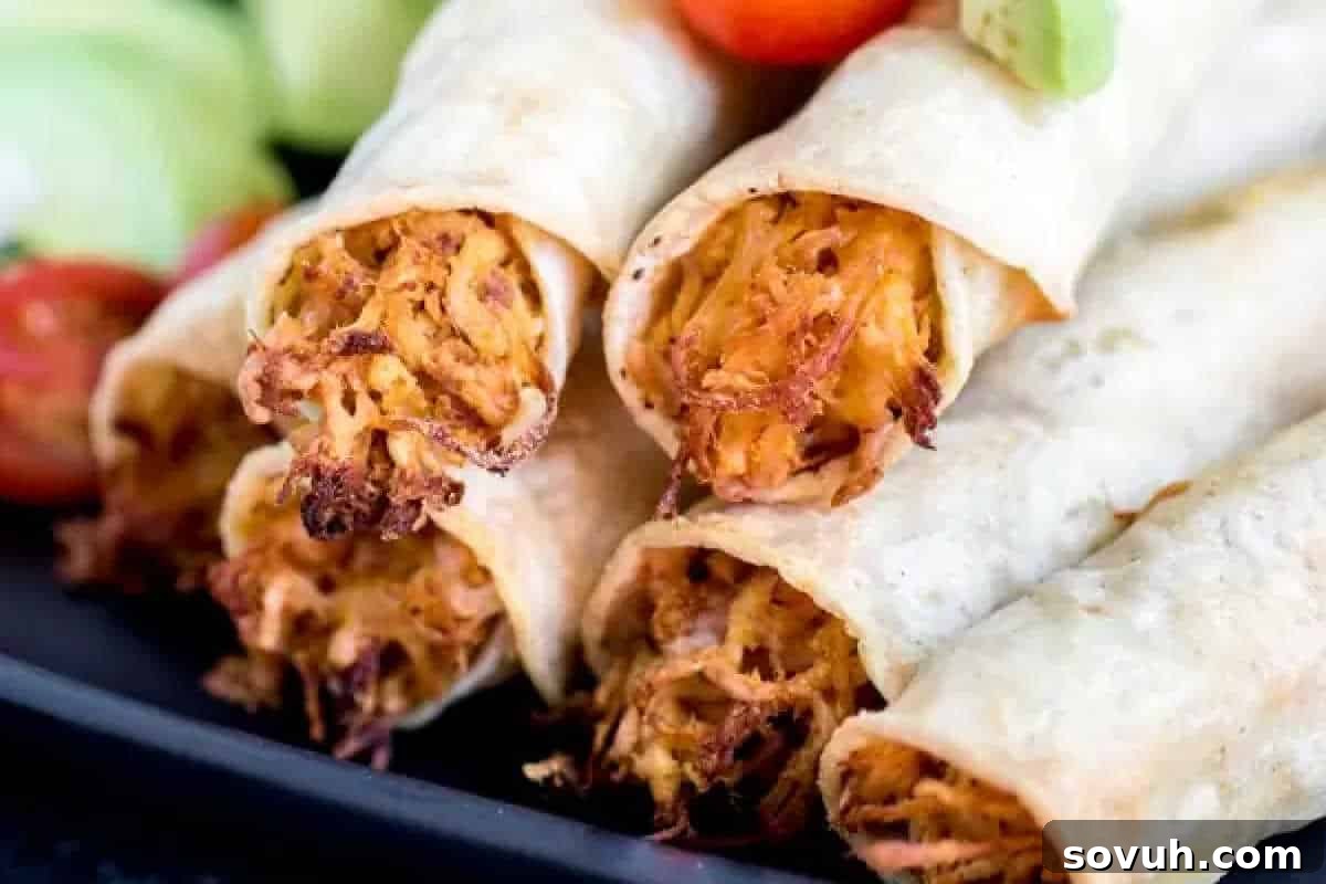 A close-up of four fried taquitos filled with shredded meat, arranged on a black plate with a side garnish of vegetables.