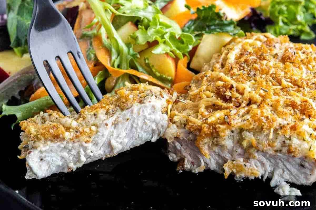 A fork piercing a slice of herb-crusted pork chop with a side of fresh mixed salad, showcasing the golden, crispy Parmesan crust.