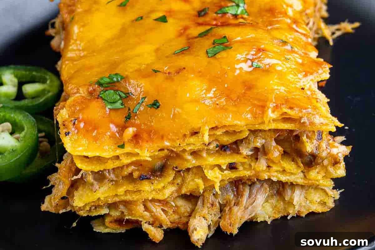 A close-up of a bubbling, layered Pulled Pork Enchilada Casserole, showcasing tender shredded meat, melted cheese, and a garnish of fresh chopped cilantro with green jalapeño slices on the side.