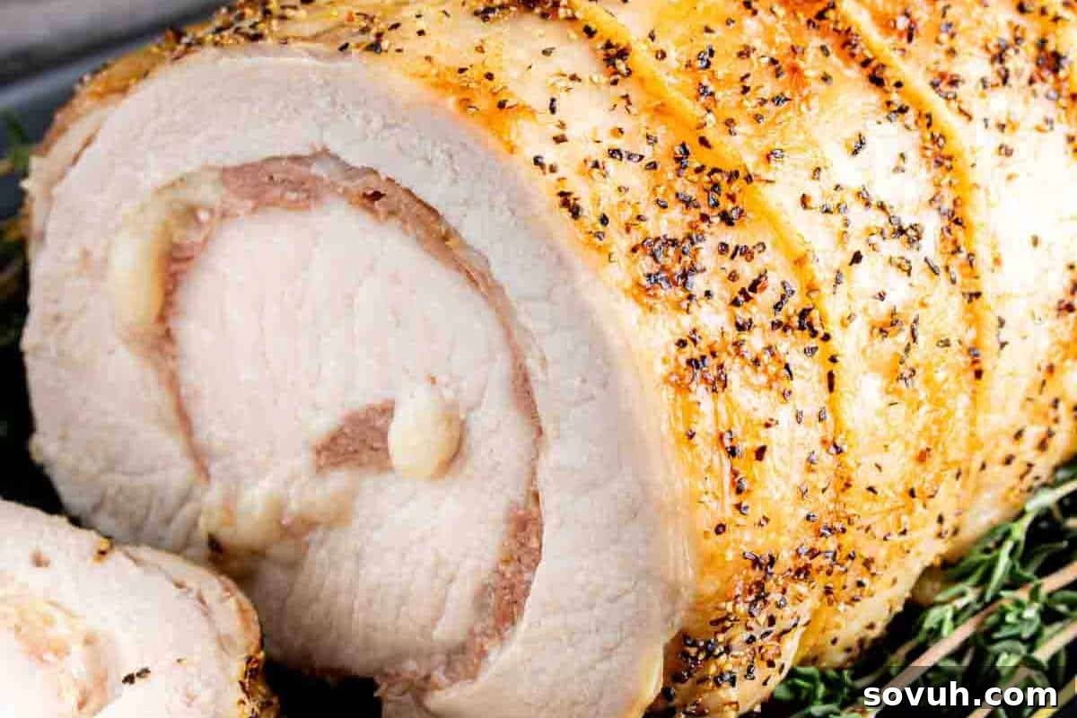 A close-up of a perfectly roasted and sliced rolled pork loin, revealing a succulent interior filled with prosciutto and provolone cheese, garnished with herbs.