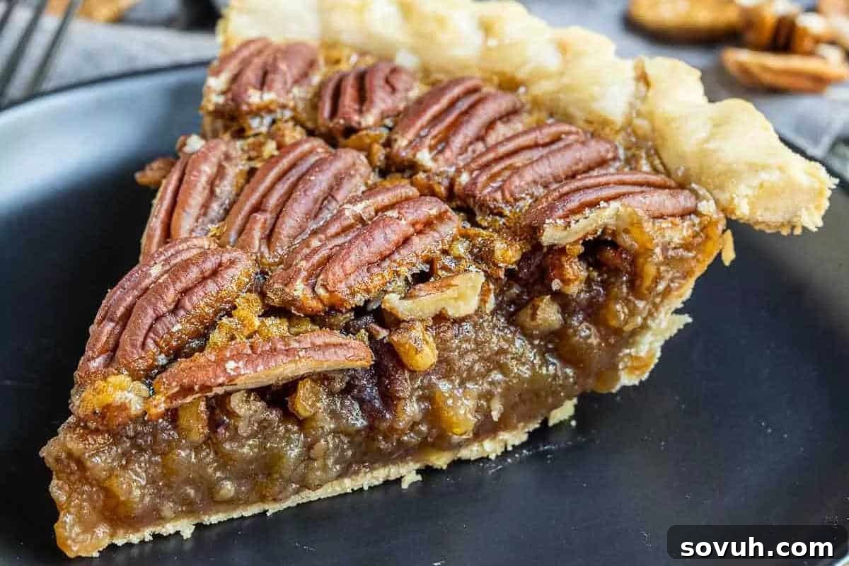 A slice of pecan pie with a layer of whole pecans on top, a gooey filling, and a light, flaky crust, presented on a black plate.