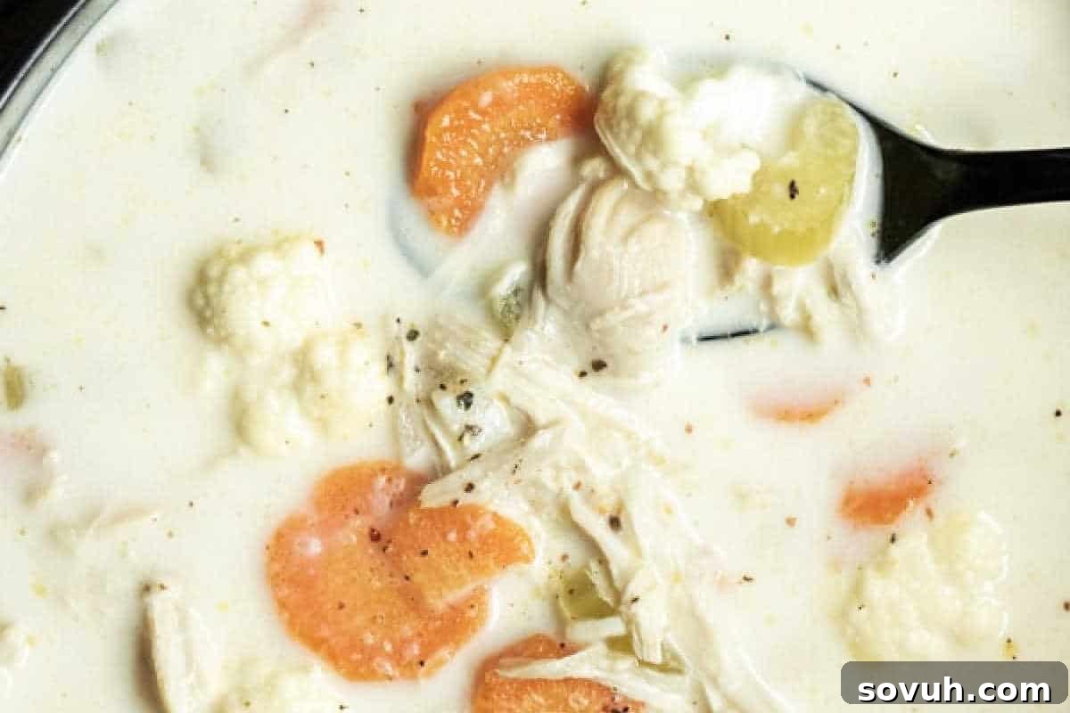 A close-up photo of a creamy soup with visible ingredients including sliced carrots, pieces of cauliflower, celery, shredded chicken, and black pepper, being stirred by a black spoon.