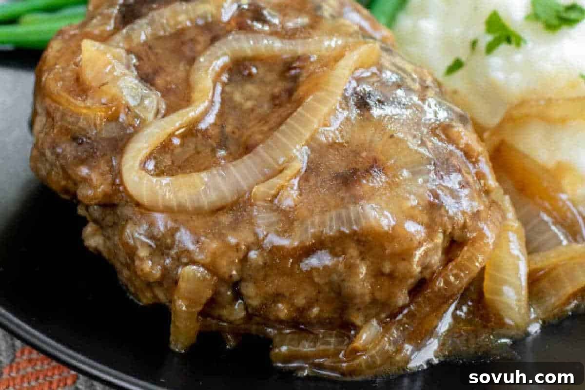 A close-up of Salisbury steak topped with caramelized onions and gravy, served with a side of mashed potatoes and green beans.
