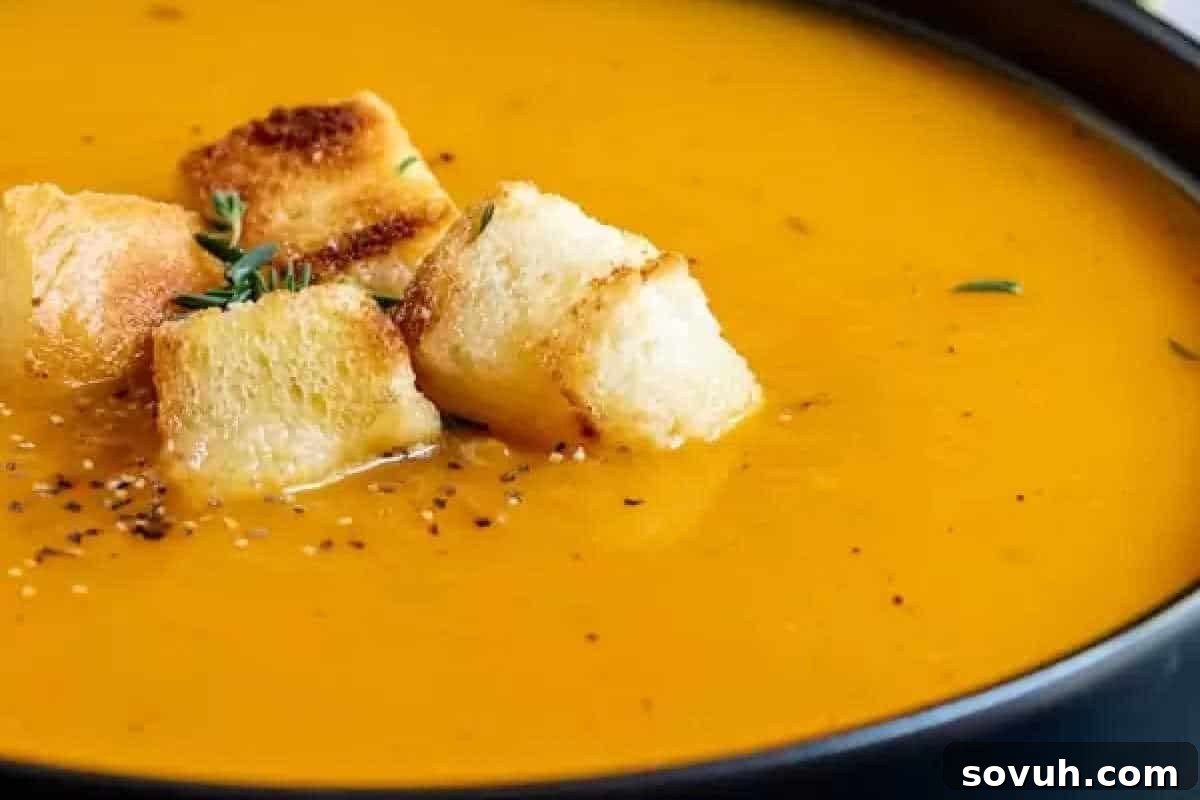 A bowl of creamy orange-colored soup, garnished with croutons and small herb leaves.