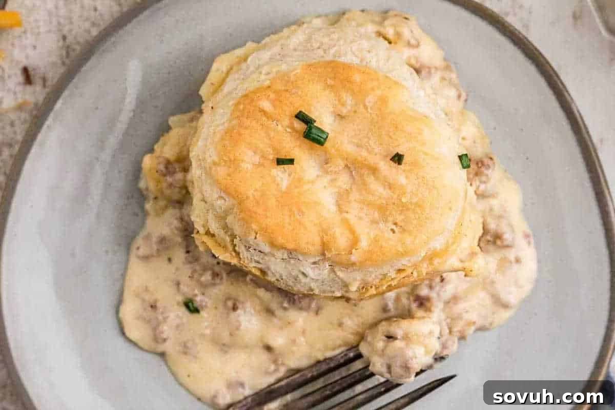 A biscuit sits on a plate covered in sausage gravy and garnished with chopped chives. A fork rests at the bottom of the plate.