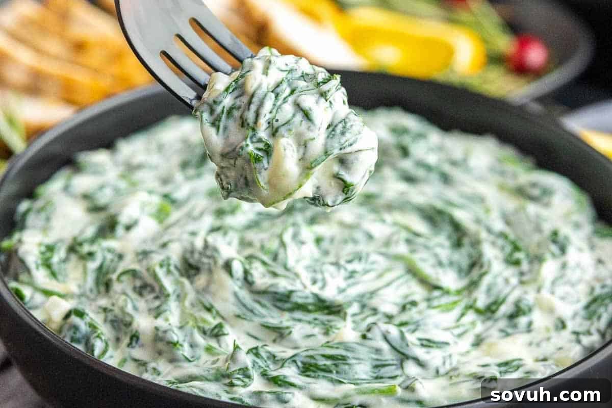 A fork holding a portion of creamy spinach dip over a black bowl filled with the same dip.
