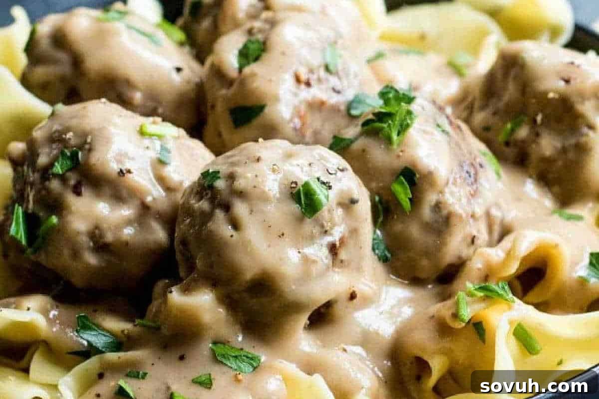 Close-up of meatballs covered in creamy sauce, garnished with chopped parsley, and served over a bed of egg noodles.
