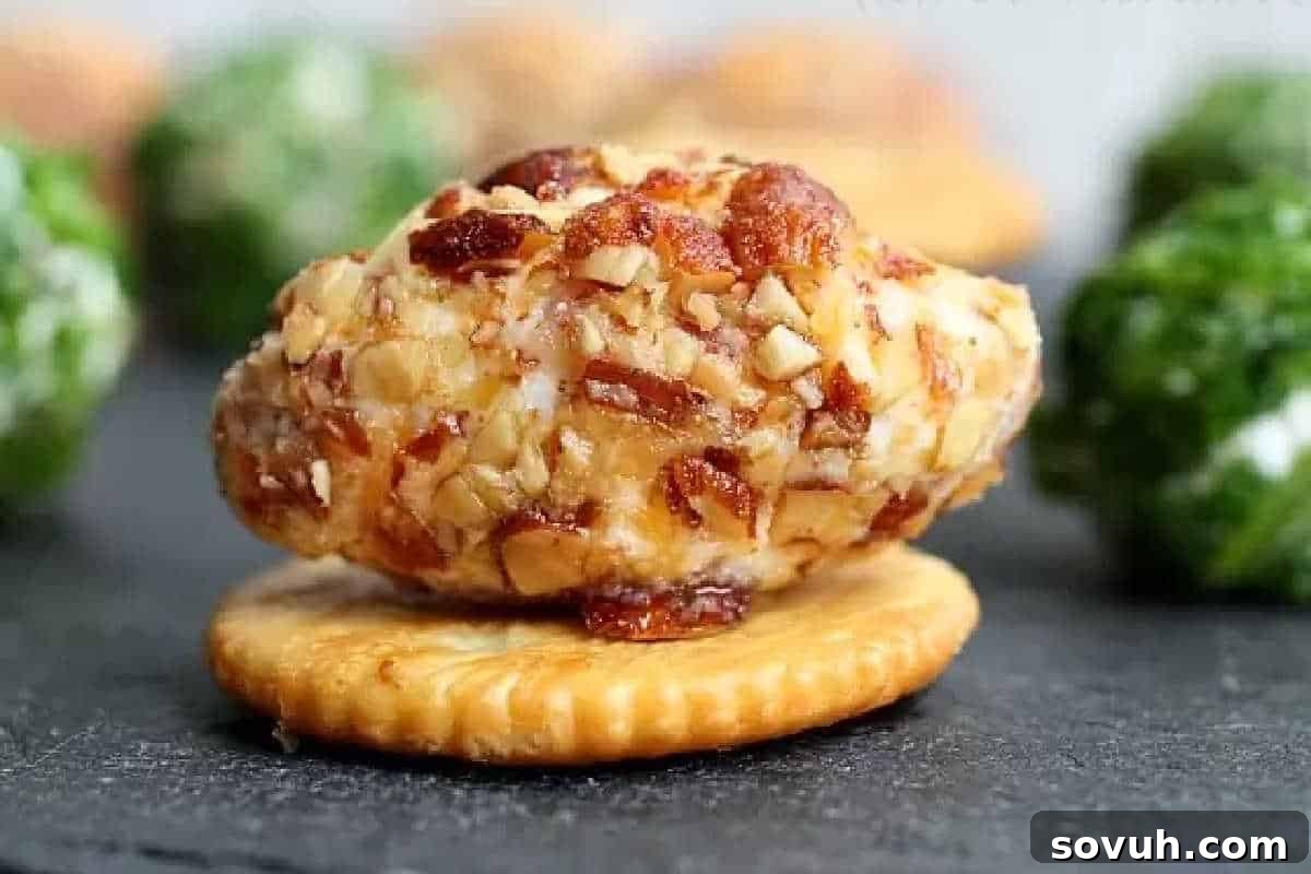 A close-up of a cheese ball coated with crushed nuts on a round cracker, placed on a dark surface with other cheese balls blurred in the background, emphasizing its delicious texture. These savory bites promise a burst of flavor, perfect for easy snacking.