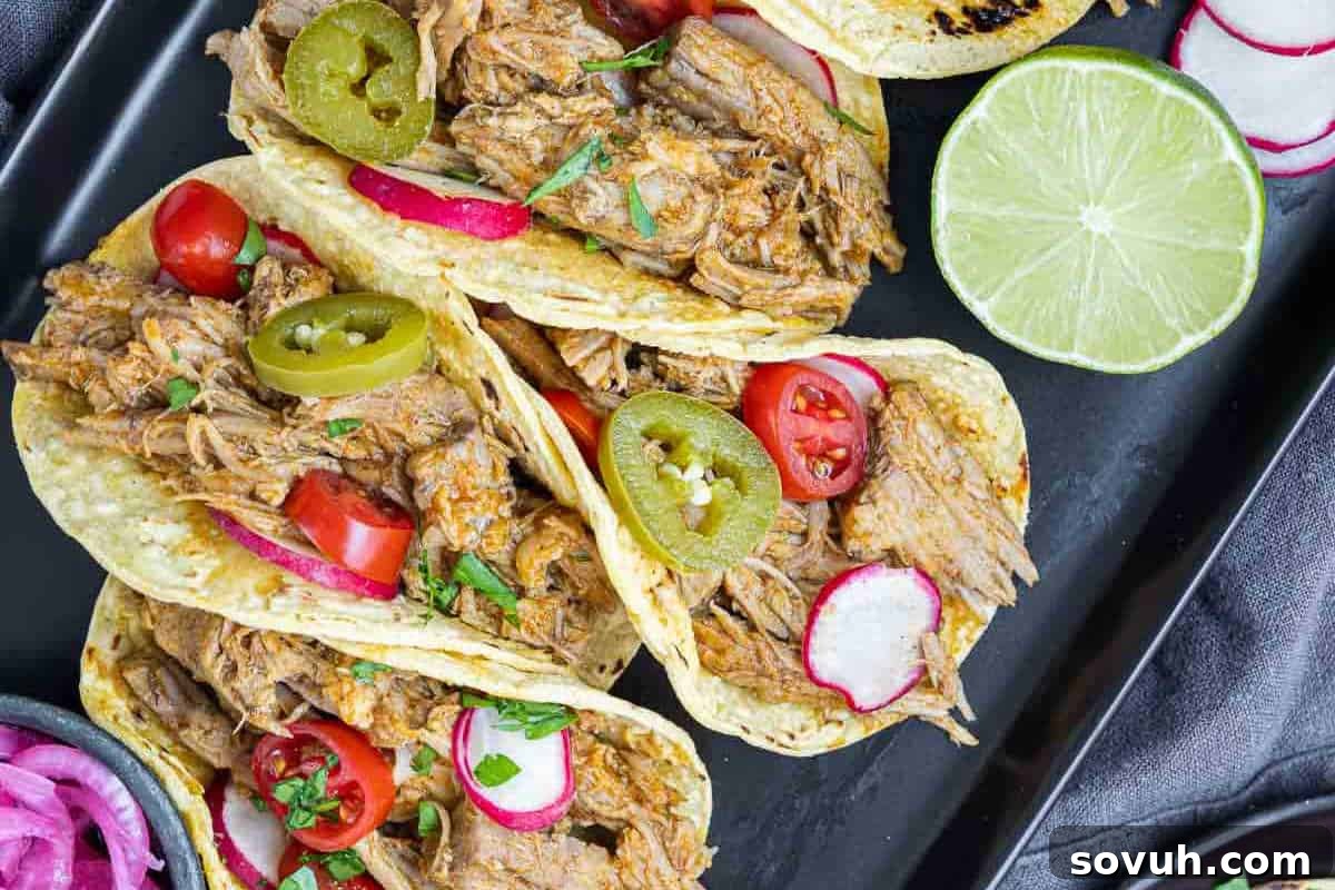 Three tacos filled with shredded meat, jalapeño slices, cherry tomatoes, radish slices, and garnished with cilantro, served on a black tray next to a halved lime, creating a colorful and inviting presentation. These tacos look fresh and ready for a satisfying meal.