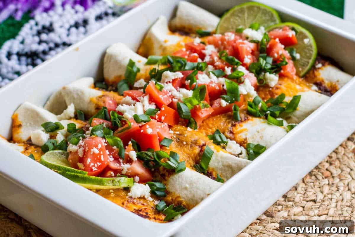 A dish of enchiladas topped with diced tomatoes, green onions, crumbled cheese, and lime wedges in a white baking dish, presenting a vibrant and appetizing meal. The enchiladas look rich and hearty, promising a delightful culinary experience.