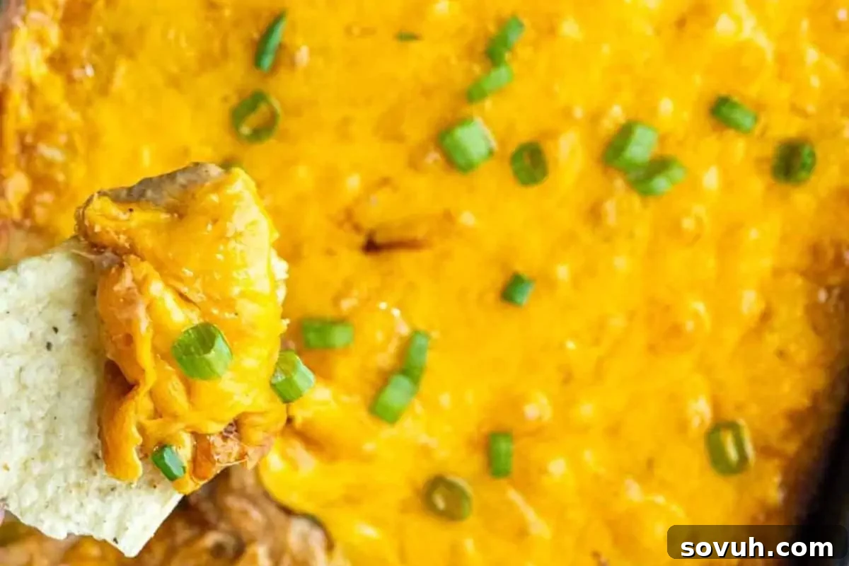 A baking dish filled with melted cheesy dip, topped with chopped green onions, with a chip scooping up some of the dip, highlighting its creamy texture. The dip looks warm and inviting, perfect for a party snack.