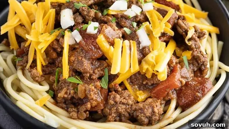 Two bowls of spaghetti topped with ground beef, shredded cheese, and diced onions. Fresh parsley, a bulb of garlic, and an onion are in the background, along with black utensils, setting a cozy, rustic scene. The chili looks rich and hearty, ideal for a comforting meal.