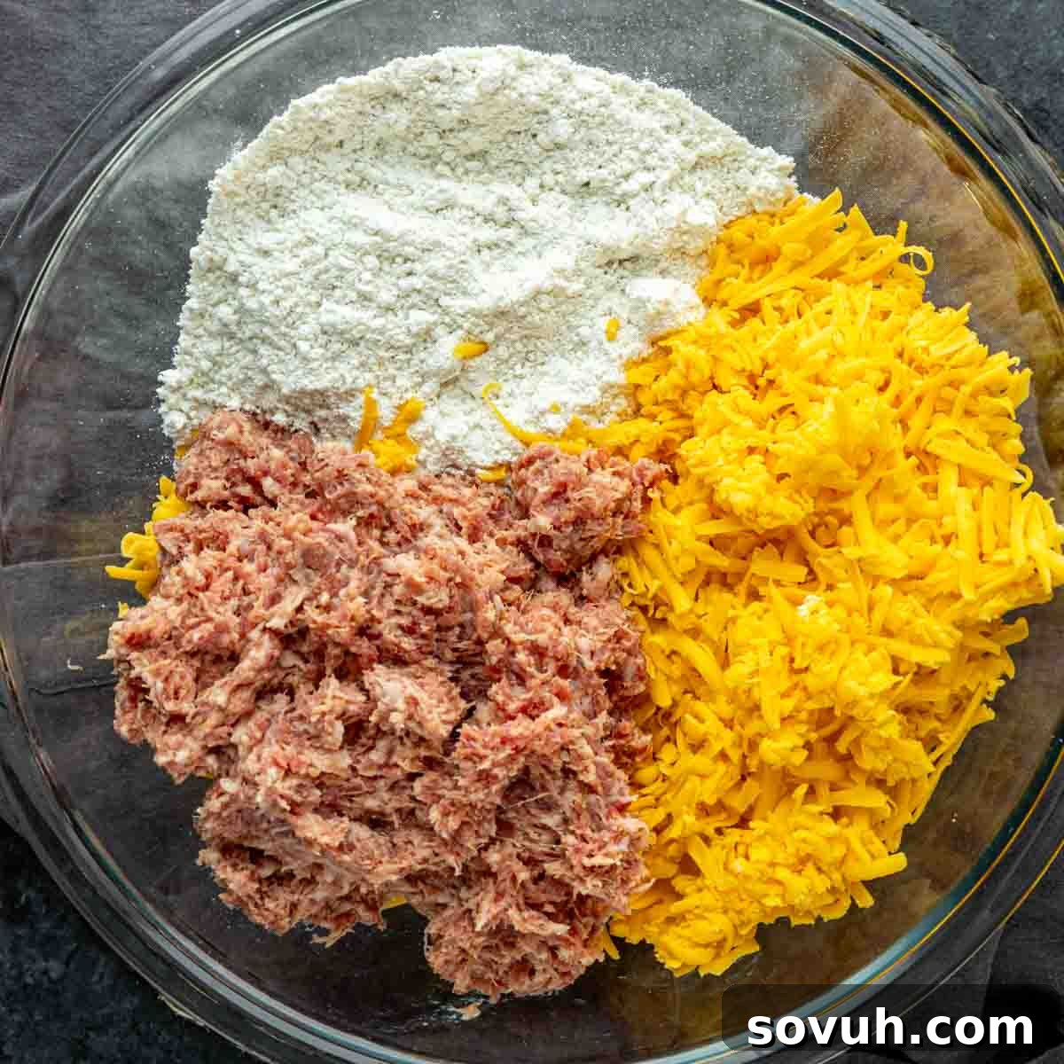 A glass bowl containing shredded cheddar cheese, flour, and ground sausage, before mixing, highlighting the raw ingredients for the sausage balls.
