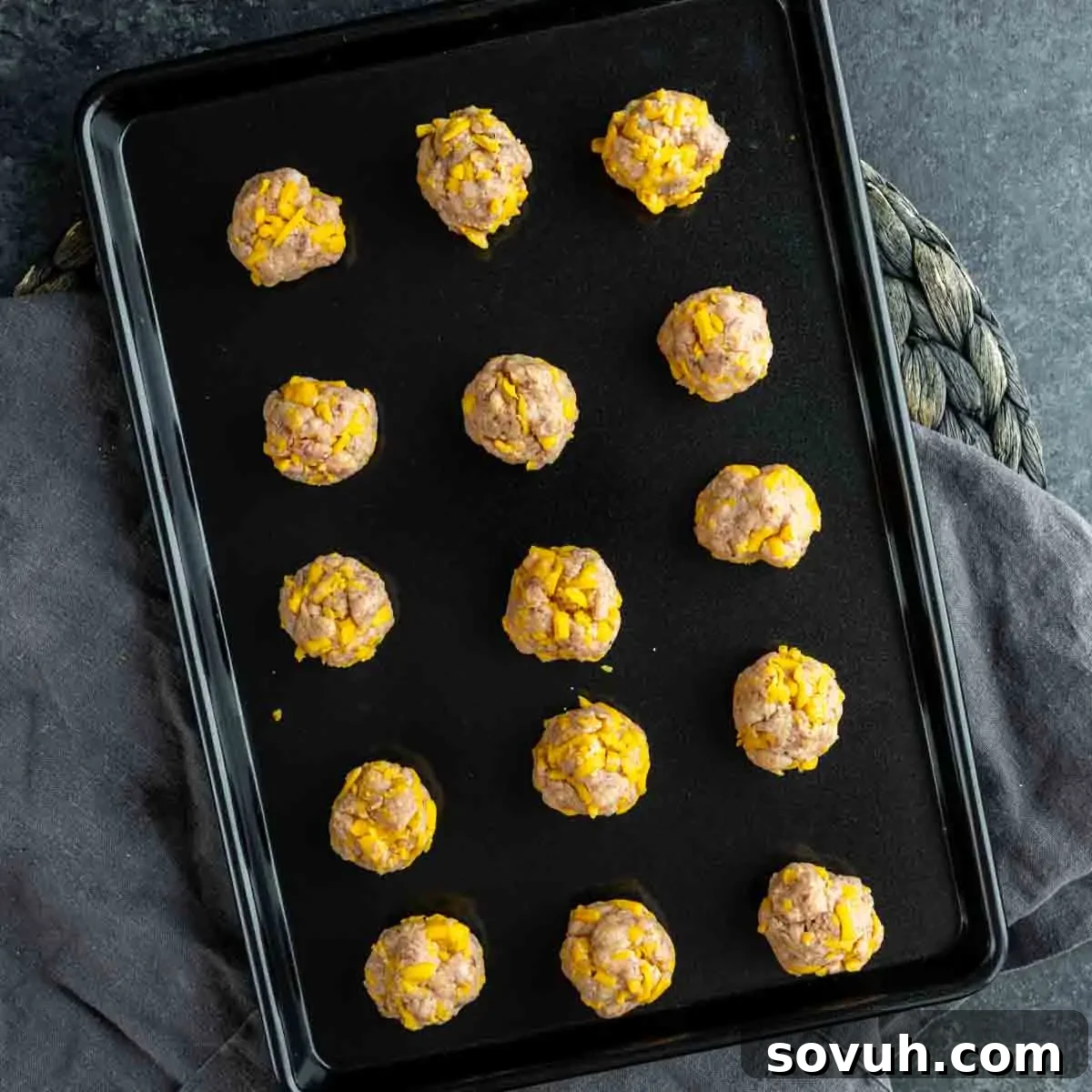 A black baking tray with 16 evenly spaced, uncooked sausage balls with visible bits of cheese, sitting on a dark surface, ready for the oven.