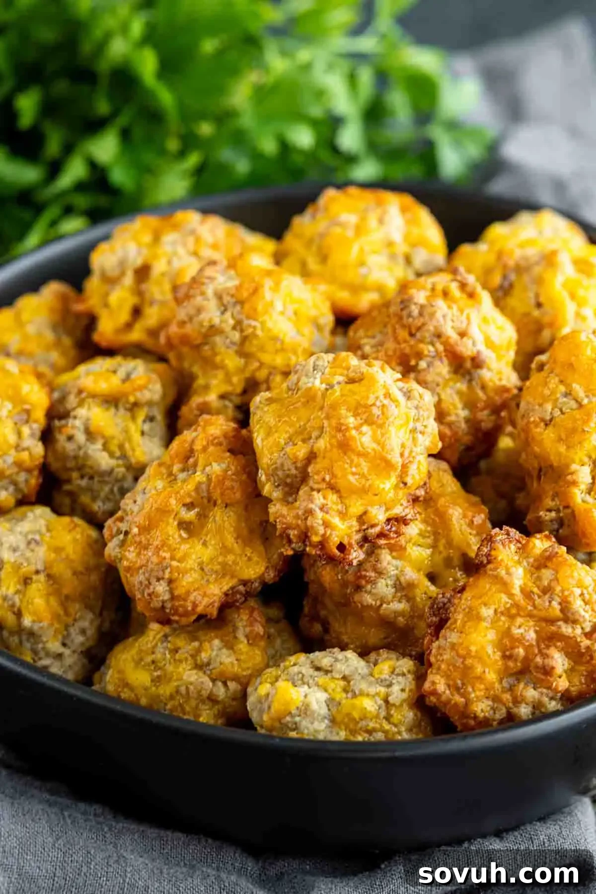 A black bowl filled with golden-brown sausage balls, placed on a gray cloth with green leafy vegetable garnish in the background, showcasing the delicious appetizer.