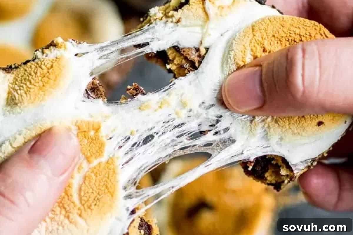Close-up of hands pulling apart a gooey dessert with toasted marshmallow layers and possibly chocolate and cookie components, showcasing a warm, melted texture—perfect for some fun baking during the cozy fall season.
