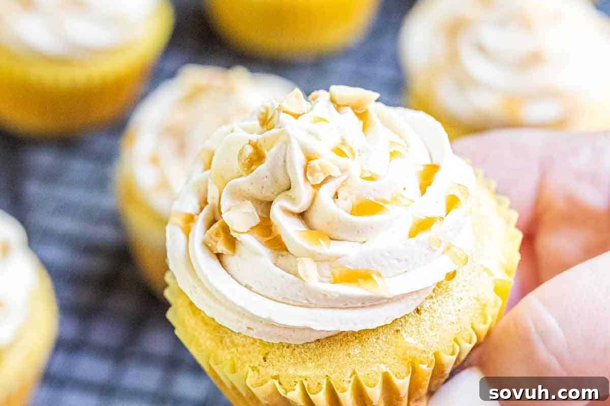 A close-up of a hand holding a vanilla cupcake topped with creamy white frosting and sprinkled with chopped nuts. Other cupcakes with similar toppings are in the background, perfect for fun baking this season.