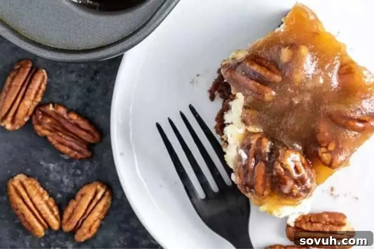 A piece of pecan pie cheesecake brownie is served on a white plate with a black fork. Several whole pecans are scattered around on the dark surface.