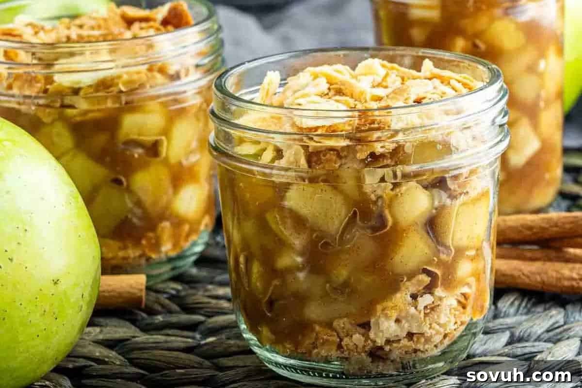 Three glass jars filled with apple crisp, placed on a woven surface with green apples and cinnamon sticks in the background.
