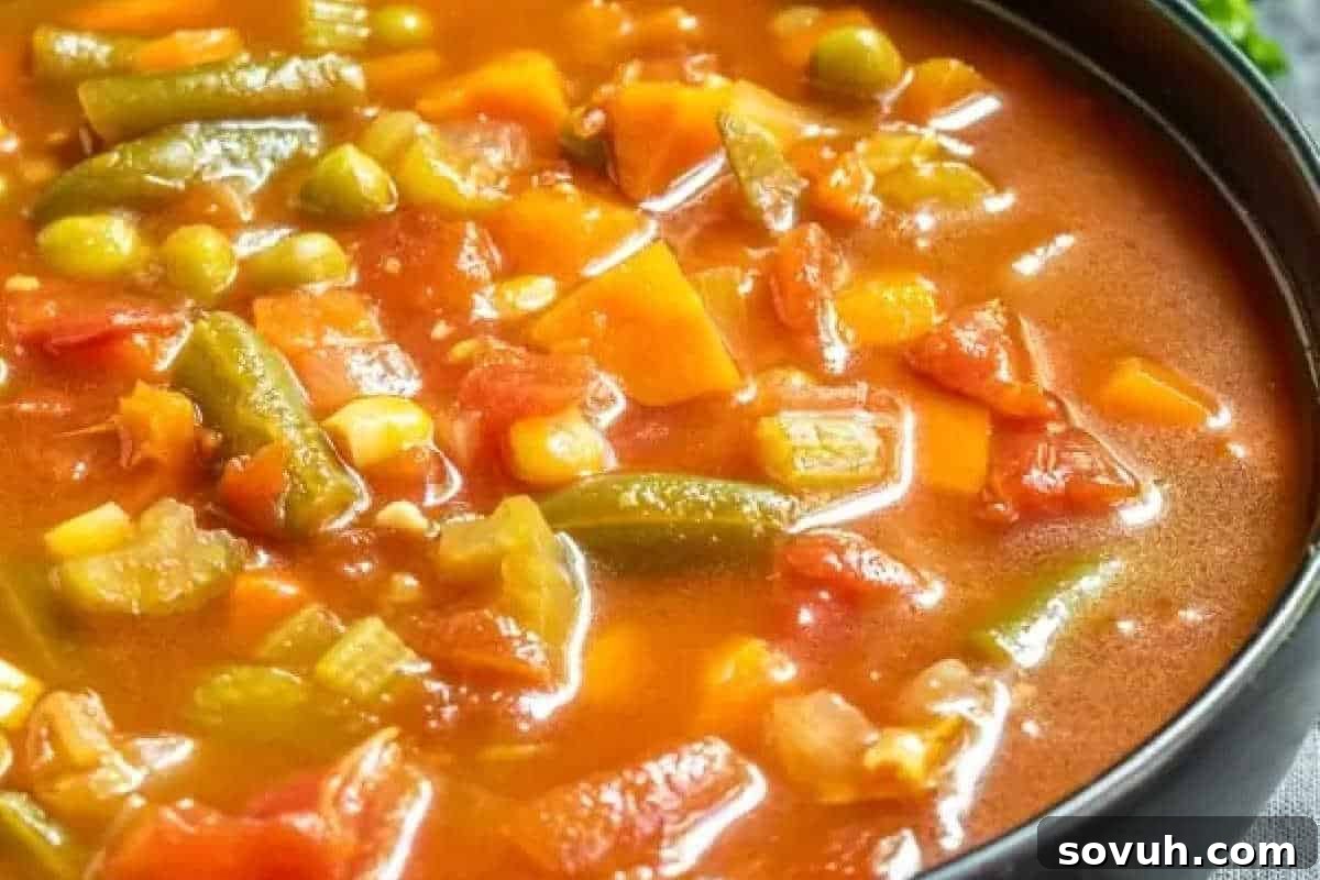 A close-up of a bowl filled with vegetable soup, featuring visible chunks of carrots, green beans, corn, peas, and tomatoes in a rich, red broth.