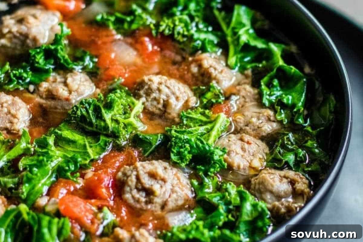 A close-up of a bowl containing a soup with meatballs, fresh kale, diced tomatoes, and broth.