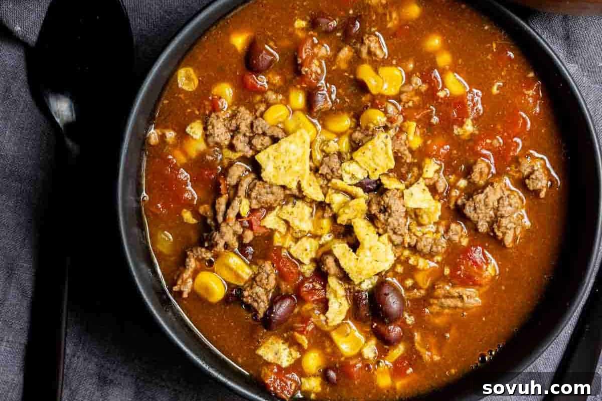 A bowl of taco soup filled with ground beef, corn, beans, tomatoes, and topped with crushed tortilla chips. A black spoon is placed to the left of the bowl.