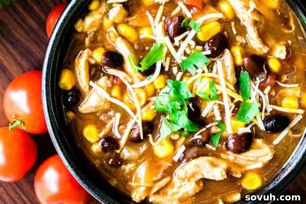 A bowl of chicken tortilla soup garnished with cilantro and shredded cheese, with visible ingredients like corn, black beans, and tomatoes. Fresh tomatoes are placed beside the bowl on a wooden surface.