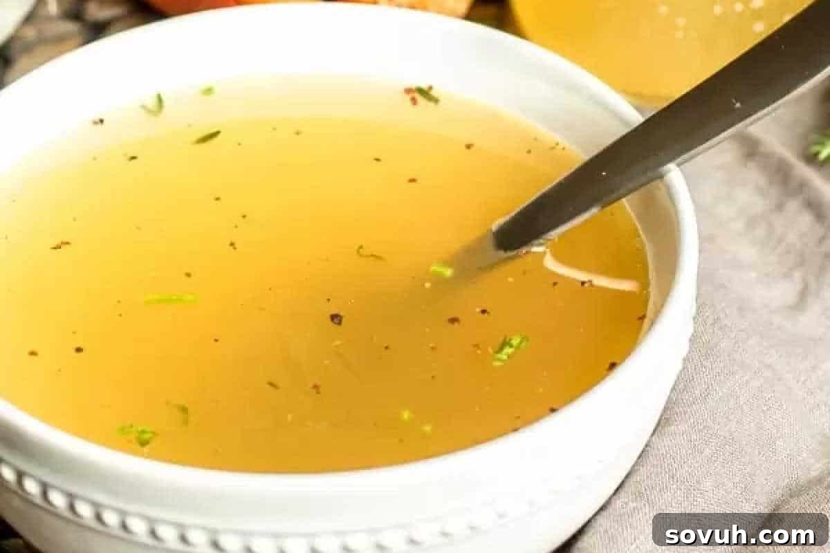 A close-up shot of a bowl of clear broth with a spoon in it. The broth contains small visible herbs and seasonings.